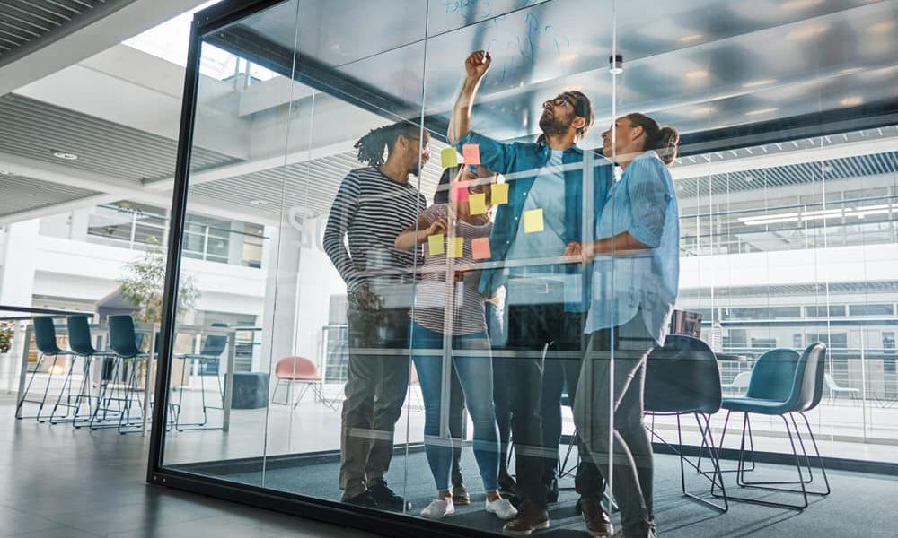 Diverse group of businesspeople drawing on the glass wall of a modern boardroom during a brainstorming session with sticky notes in an office